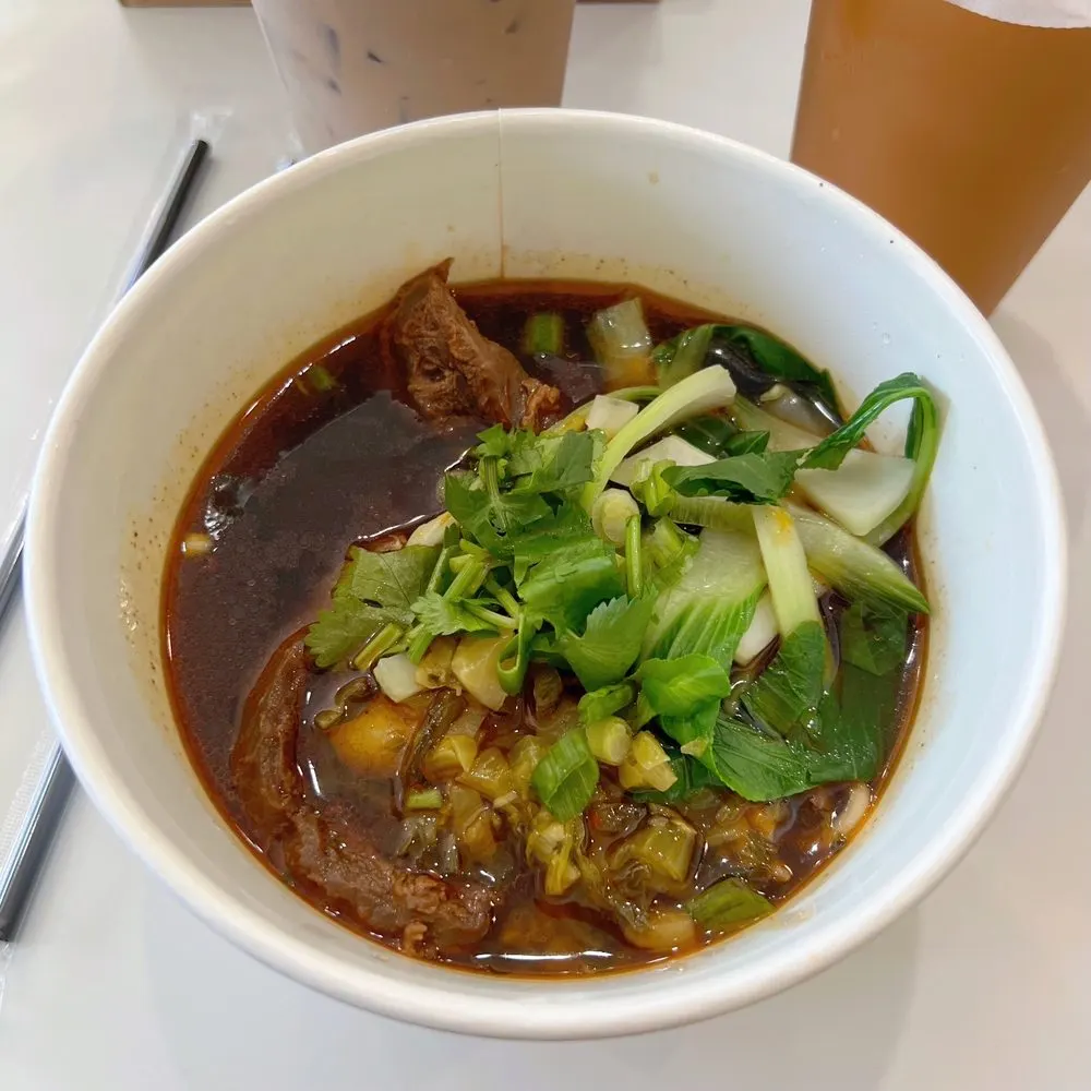 Bowl of braised beef noodle soup with bok choy, cilantro, scallions, and pickled mustard greens, with iced drinks behind it